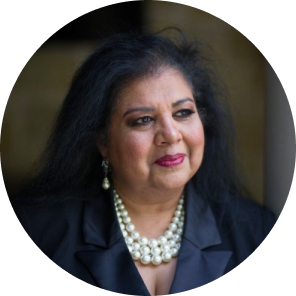 Portrait of a woman with long dark hair, wearing a black blazer and a pearl necklace, looking thoughtfully off to the side, set against a softly blurred background.