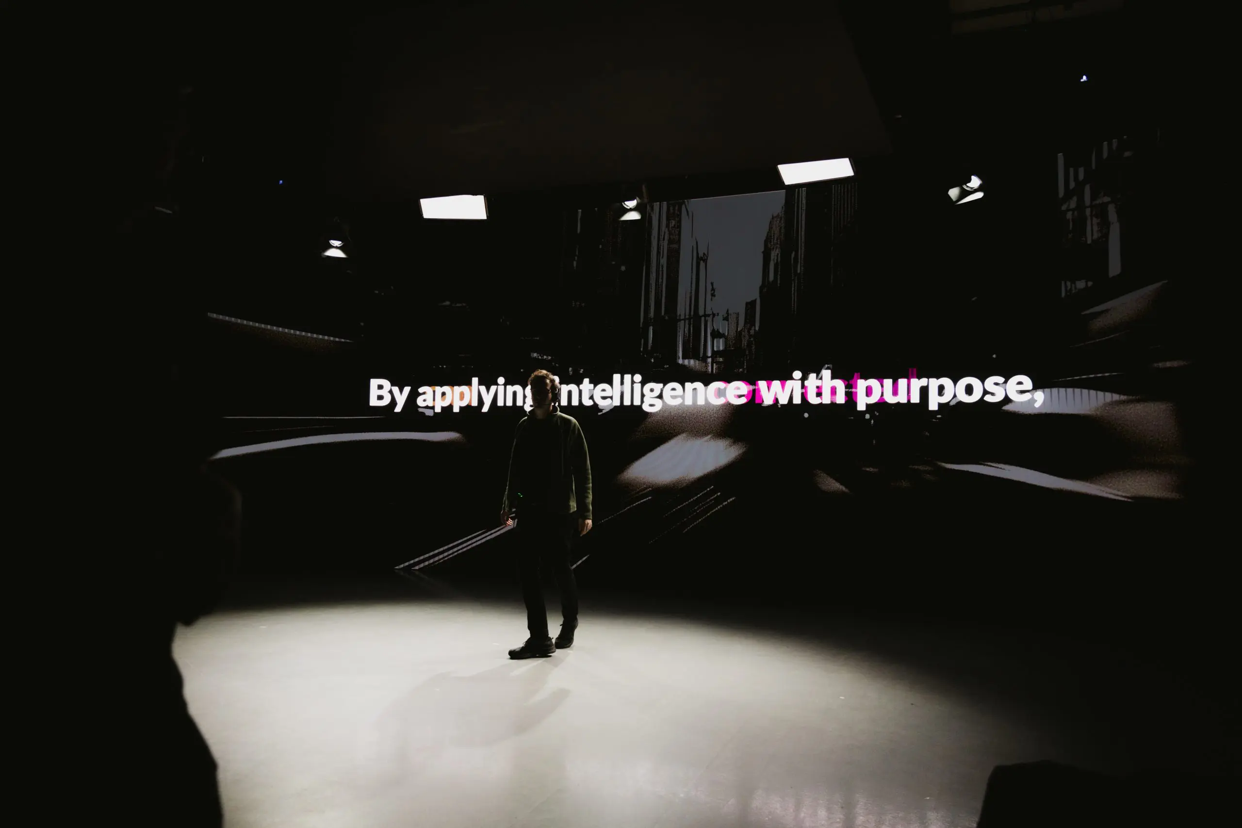 A person standing on a stage in front of a large screen displaying the message about applying intelligence with purpose, in a modern and dynamic setting.