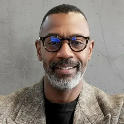 Professional portrait of a smiling middle-aged man with glasses, featuring a well-groomed beard and short hair, dressed in a stylish blazer against a textured gray background.