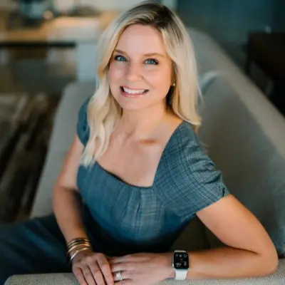 Smiling woman with long blonde hair wearing a gray dress, sitting on a sofa, showcasing a modern interior and an Apple Watch on her wrist.