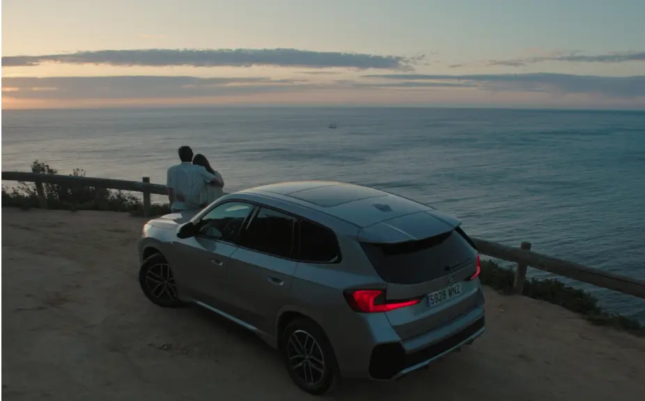 Couple enjoying a romantic sunset view by the ocean with a silver SUV parked nearby, highlighting a scenic coastal landscape.