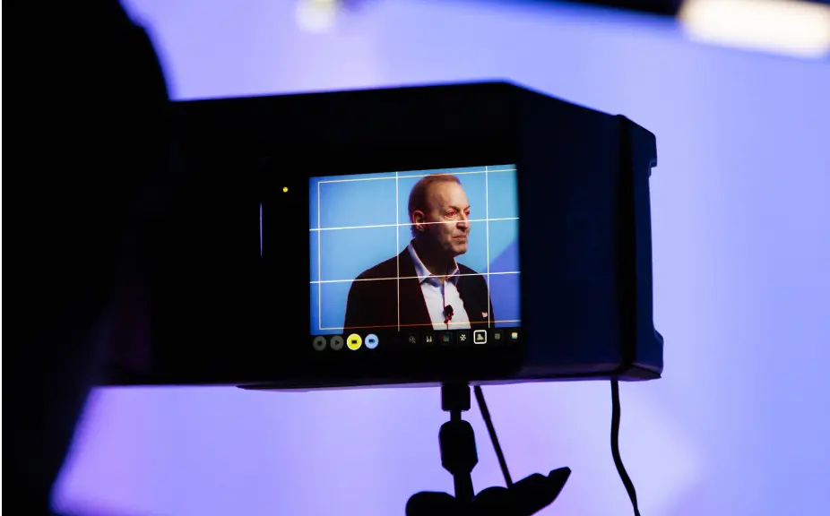 Camera viewfinder capturing a speaker during a presentation, with a blue background and grid overlay for composition. The focus is on the speaker's profile, highlighting the event's professional setting.