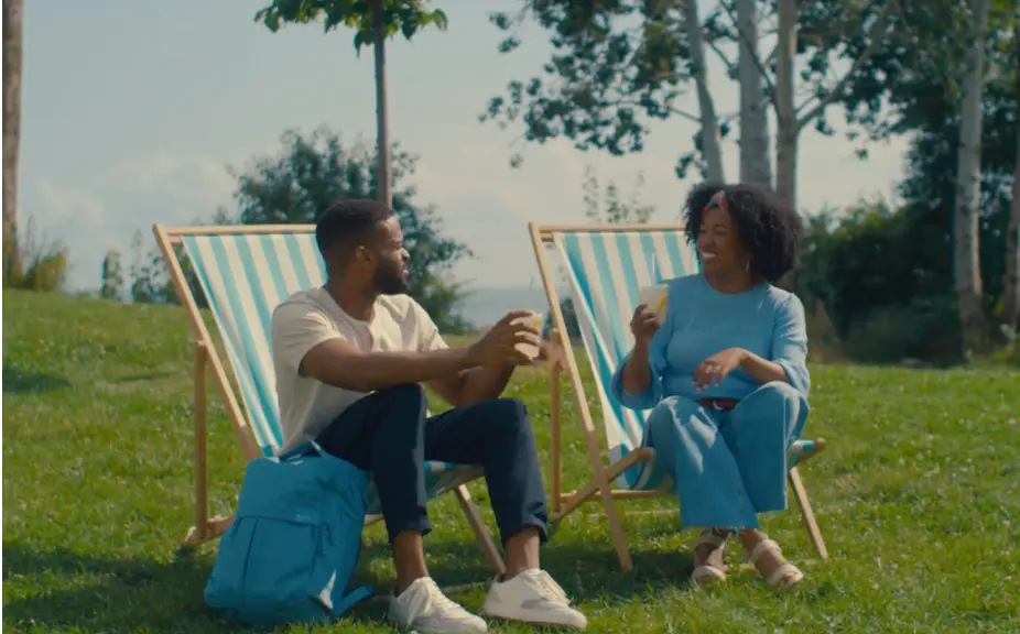 Two friends enjoying drinks while seated on deck chairs in a sunny park, surrounded by greenery and trees, creating a relaxed outdoor atmosphere.
