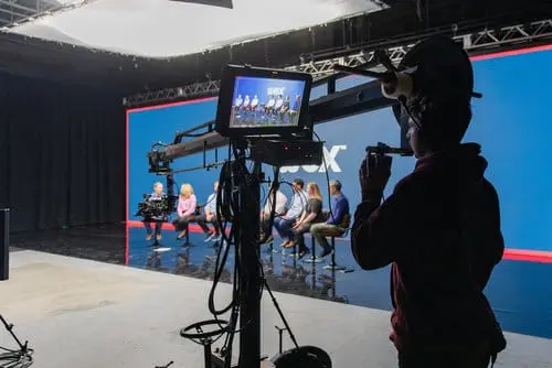 A camera operator using a crane to film a panel discussion in a studio setting, with a blue backdrop and a group of speakers seated on stage.