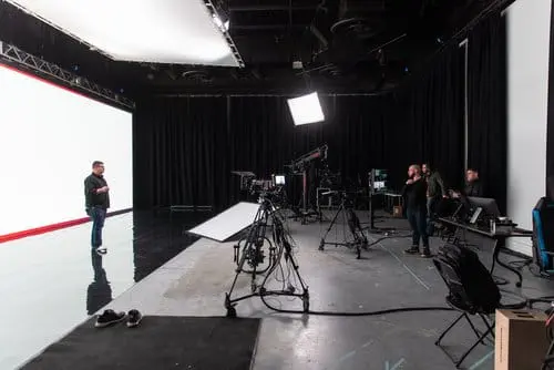 Film studio interior featuring a large empty backdrop, production crew members, and various camera equipment. The space is equipped for video shoots, with lighting rigs and monitors visible, showcasing a professional filming environment.