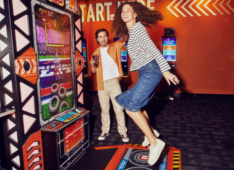 Young adults enjoying a fun moment at an arcade, playing an interactive sports game. The woman is jumping in excitement, while the man smiles and holds a drink. The vibrant arcade environment features colorful screens and game machines.