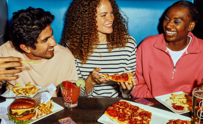 Group of friends enjoying a meal at a restaurant, featuring a hamburger, fries, and pizza. They are laughing and sharing food, highlighting a fun dining experience.