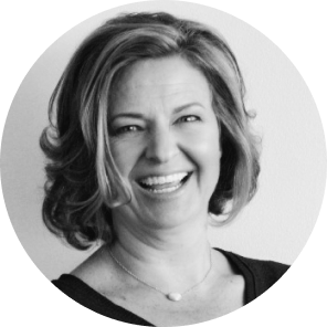 Smiling woman with shoulder-length curly hair, wearing a black top and a simple necklace, captured in a circular portrait against a light background.