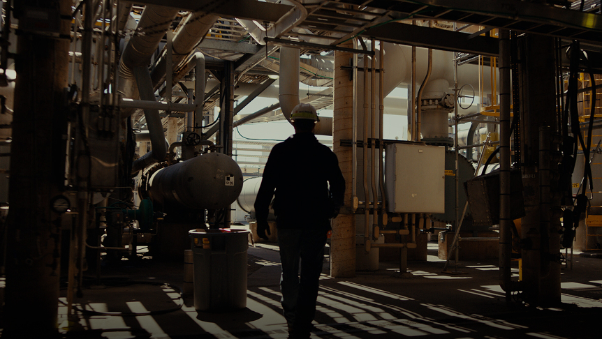 Worker in a hard hat walking through a complex industrial facility with pipes and machinery, highlighting the operational environment of manufacturing or processing industries.