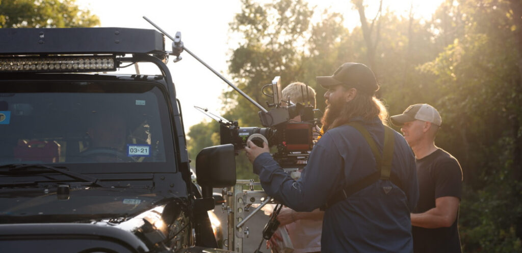 Film crew capturing footage with a camera mounted on a support rig, set near a black vehicle in a natural outdoor environment during golden hour.