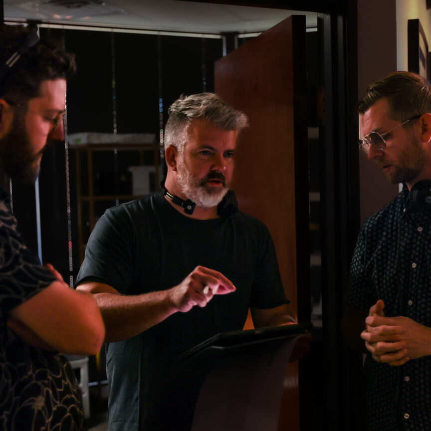 Three professionals engaged in a discussion, with one man pointing at a tablet while the others listen attentively. The setting appears to be a modern office or studio, highlighted by low ambient lighting.