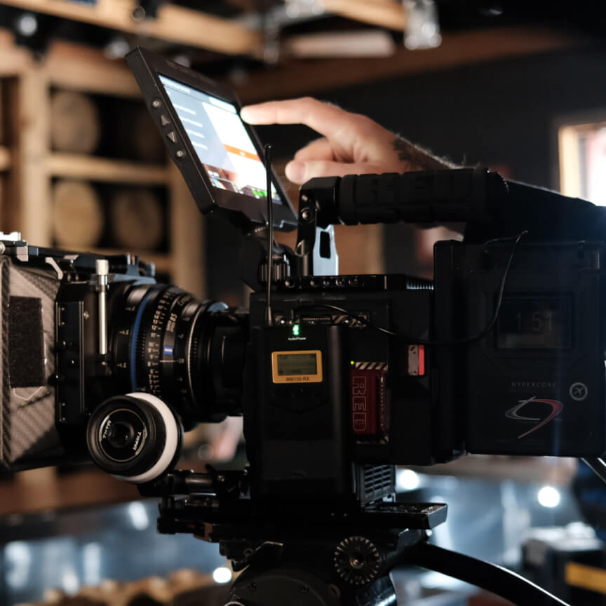 Close-up of a professional camera setup featuring a RED digital cinema camera with a focus on the touchscreen monitor being adjusted by a hand. The camera is equipped with a high-quality lens, mounted on a sturdy tripod, in a dimly lit studio environment.