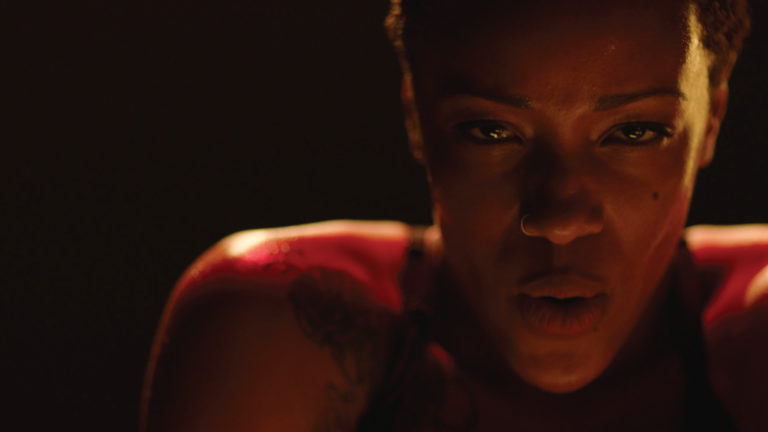 Close-up of a focused woman with tattoos, showcasing determination and intensity in a dimly lit environment, emphasizing her expressions and strength.
