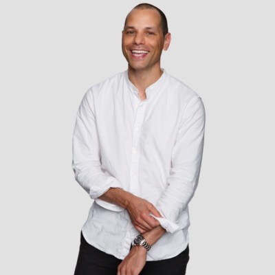Smiling man in a white button-up shirt with rolled sleeves, standing confidently with arms crossed, against a neutral gray background.