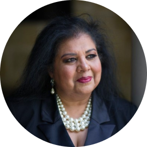 Portrait of a woman with long dark hair, wearing a black blazer and a pearl necklace, looking thoughtfully off to the side, set against a softly blurred background.