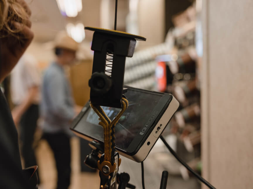 Close-up of a smartphone mounted on a stabilizing rig, showcasing a camera setup in a busy environment, with blurred figures in the background. The image highlights the use of technology in content creation or filming.