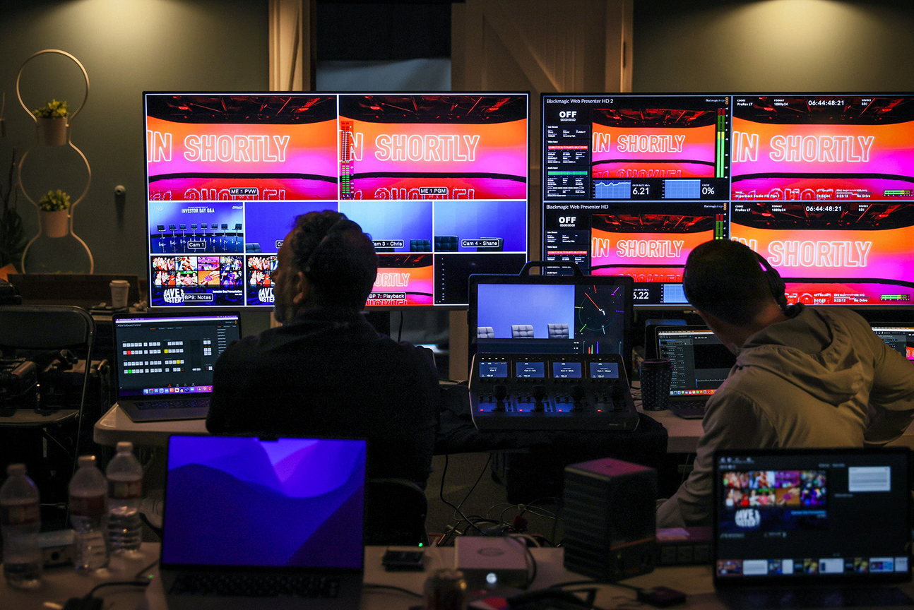Behind-the-scenes view of a live production control room featuring multiple screens displaying video feeds and graphics, with two operators managing the broadcast setup using laptops and control panels.