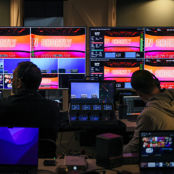 Broadcast control room with multiple screens displaying "IN SHORTLY," featuring technicians managing live video production equipment and monitoring feeds.