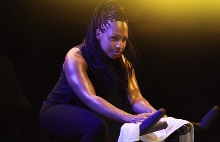 Woman with braided hair exercising on a stationary bike, focused and determined, with dramatic lighting highlighting her muscles in a fitness studio.
