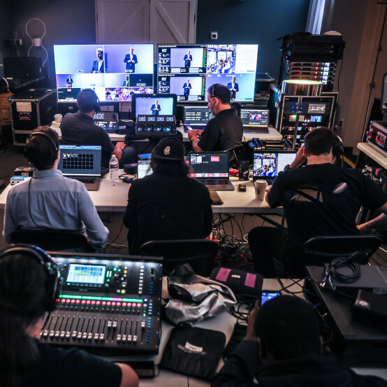 A group of technicians operating video production equipment in a control room, with multiple screens displaying a live presentation, audio mixing consoles, and laptops, highlighting a collaborative environment for event broadcasting.