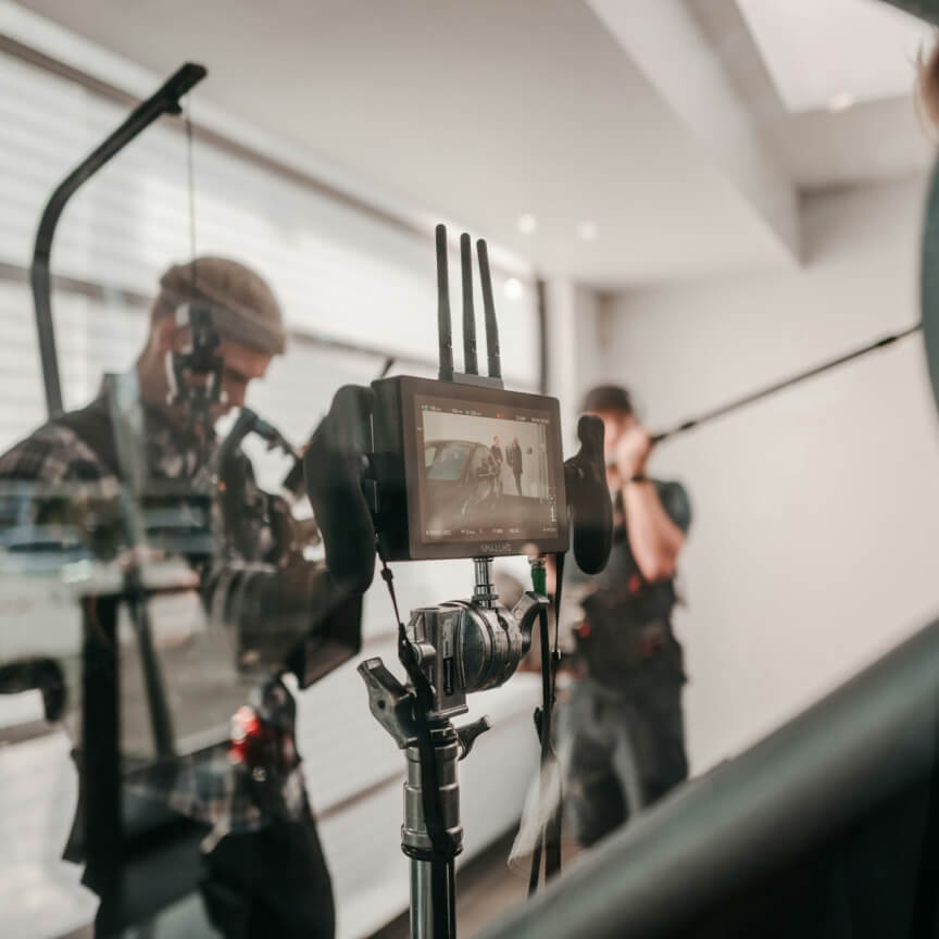 Behind-the-scenes view of a film set featuring a camera setup with a monitor displaying a scene being filmed, while crew members operate equipment in a well-lit studio environment.