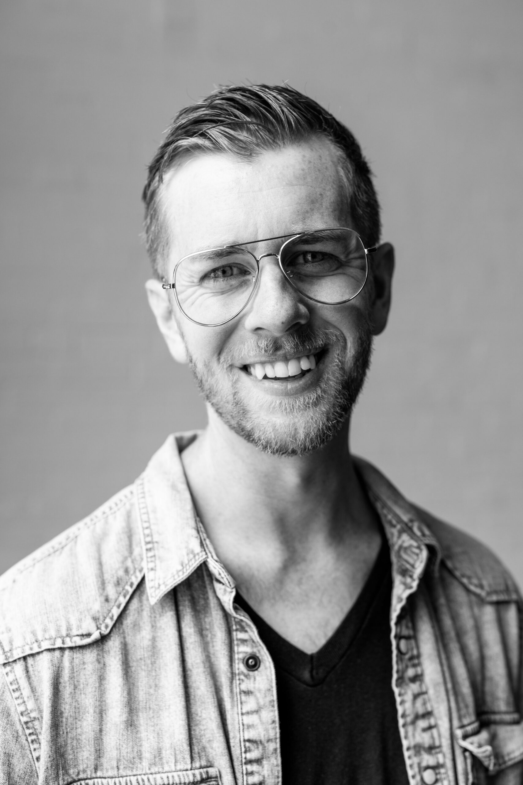 Smiling man with glasses wearing a denim jacket over a black shirt, posing against a light gray background.