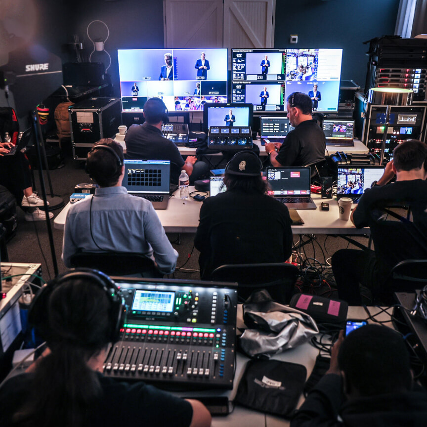 Control room with multiple monitors and audio equipment, showcasing a live event production setup with technicians managing video feeds and sound.