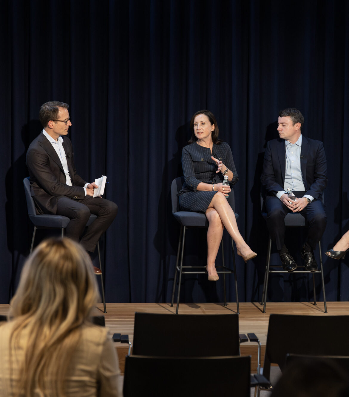 Panel discussion featuring four speakers, including a moderator and three panelists, seated on high stools against a dark blue backdrop. The audience is visible in the foreground, focusing on the speakers. The setting suggests a professional event centered on industry insights and expert opinions.