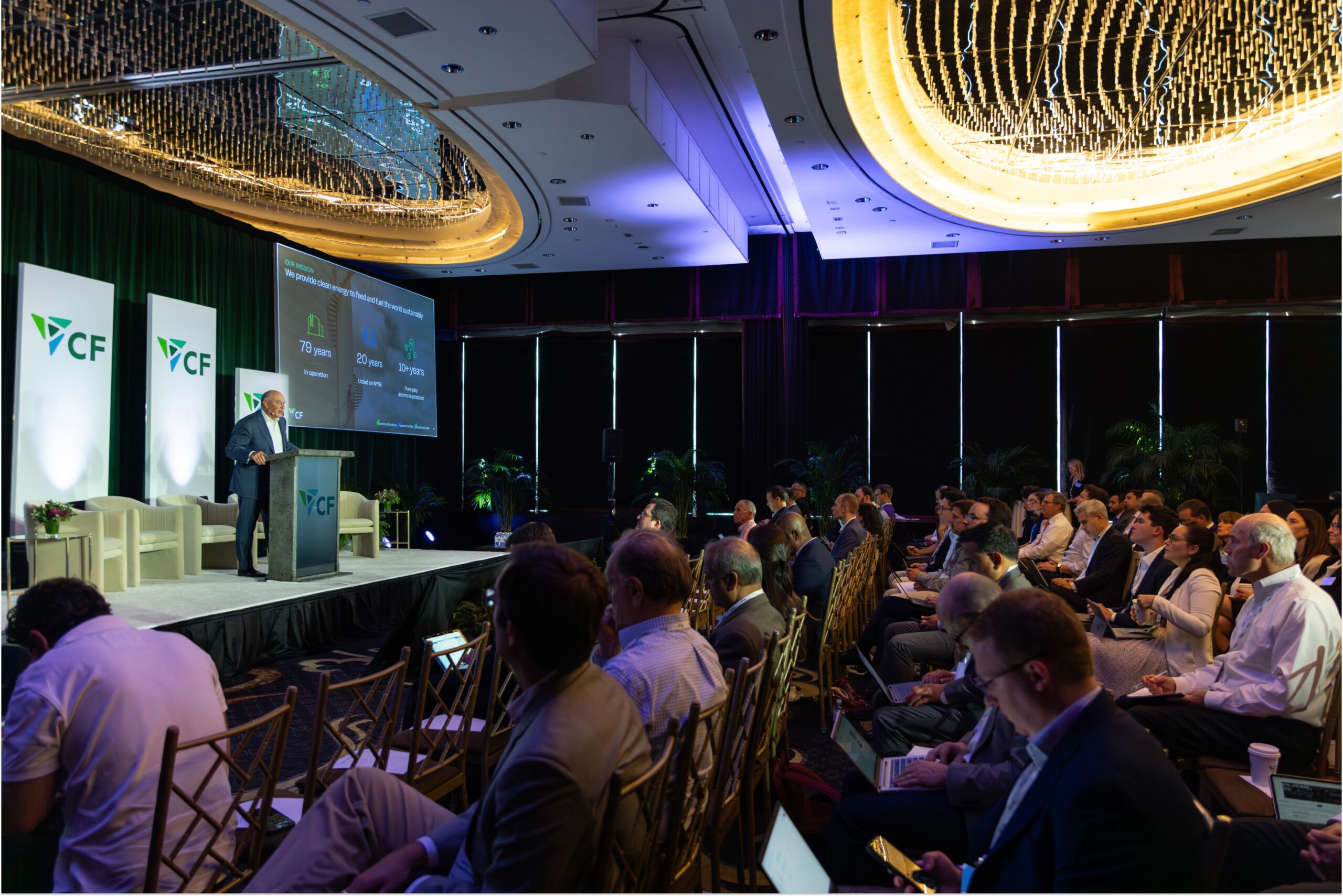 Conference speaker presenting on stage at a corporate event, with an audience engaged in the foreground. The backdrop features the CF logo and a presentation screen highlighting key statistics related to the organization's history and accomplishments. Elegant chandeliers illuminate the venue, enhancing the professional atmosphere.