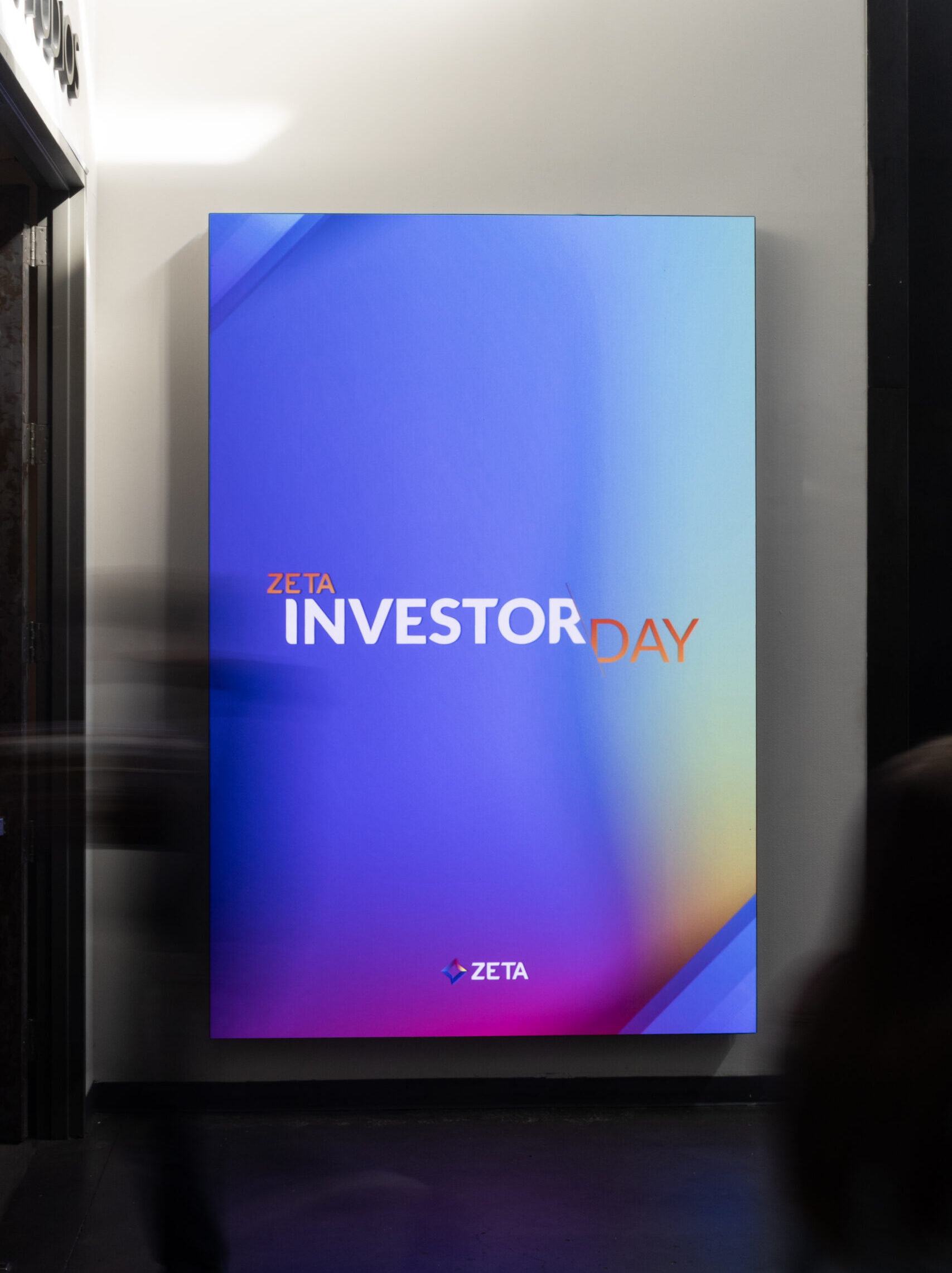 Zeta Investor Day promotional display featuring a vibrant blue and purple gradient background with the text "ZETA INVESTOR DAY" prominently displayed. The image captures attendees in motion, emphasizing the event's dynamic atmosphere.