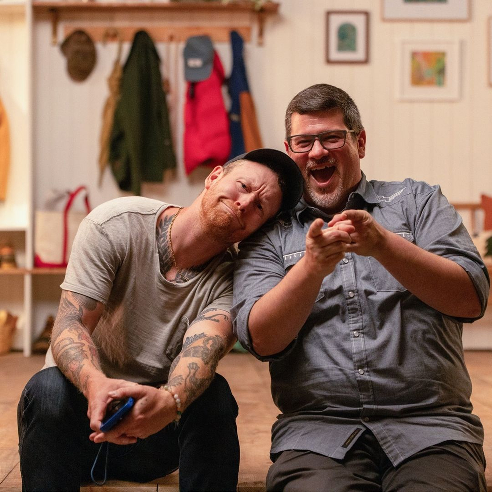 Two friends sitting together indoors, displaying playful expressions and laughter. One has tattoos and a cap, while the other wears glasses and a button-up shirt. The background features a cozy setting with hanging clothing and framed artwork, creating a warm atmosphere.