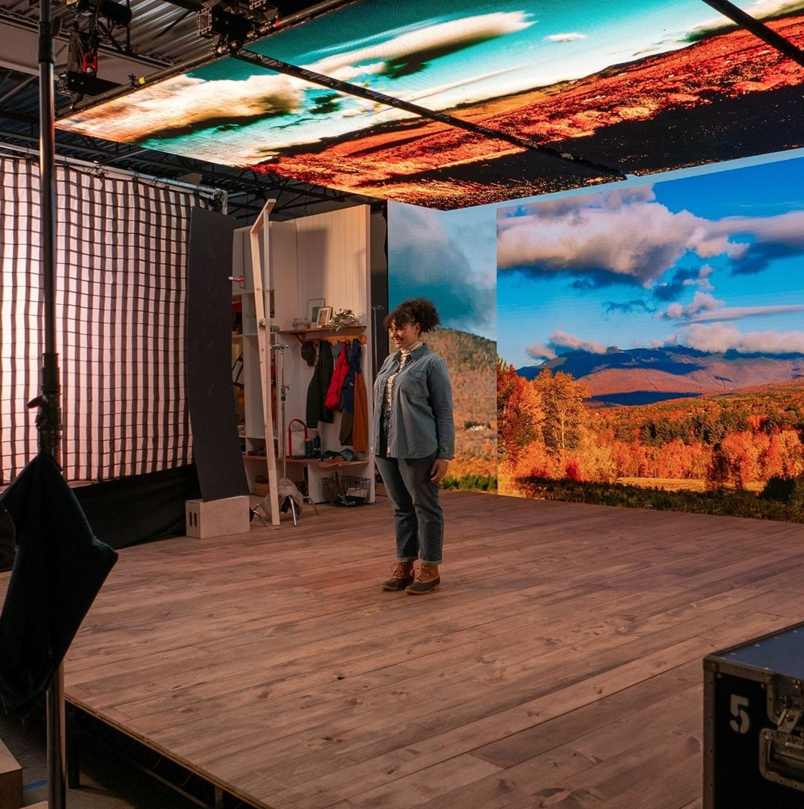 Woman standing on a wooden stage in a photography studio, surrounded by vibrant digital backdrops of autumn landscapes and a colorful ceiling display, showcasing a creative workspace environment.