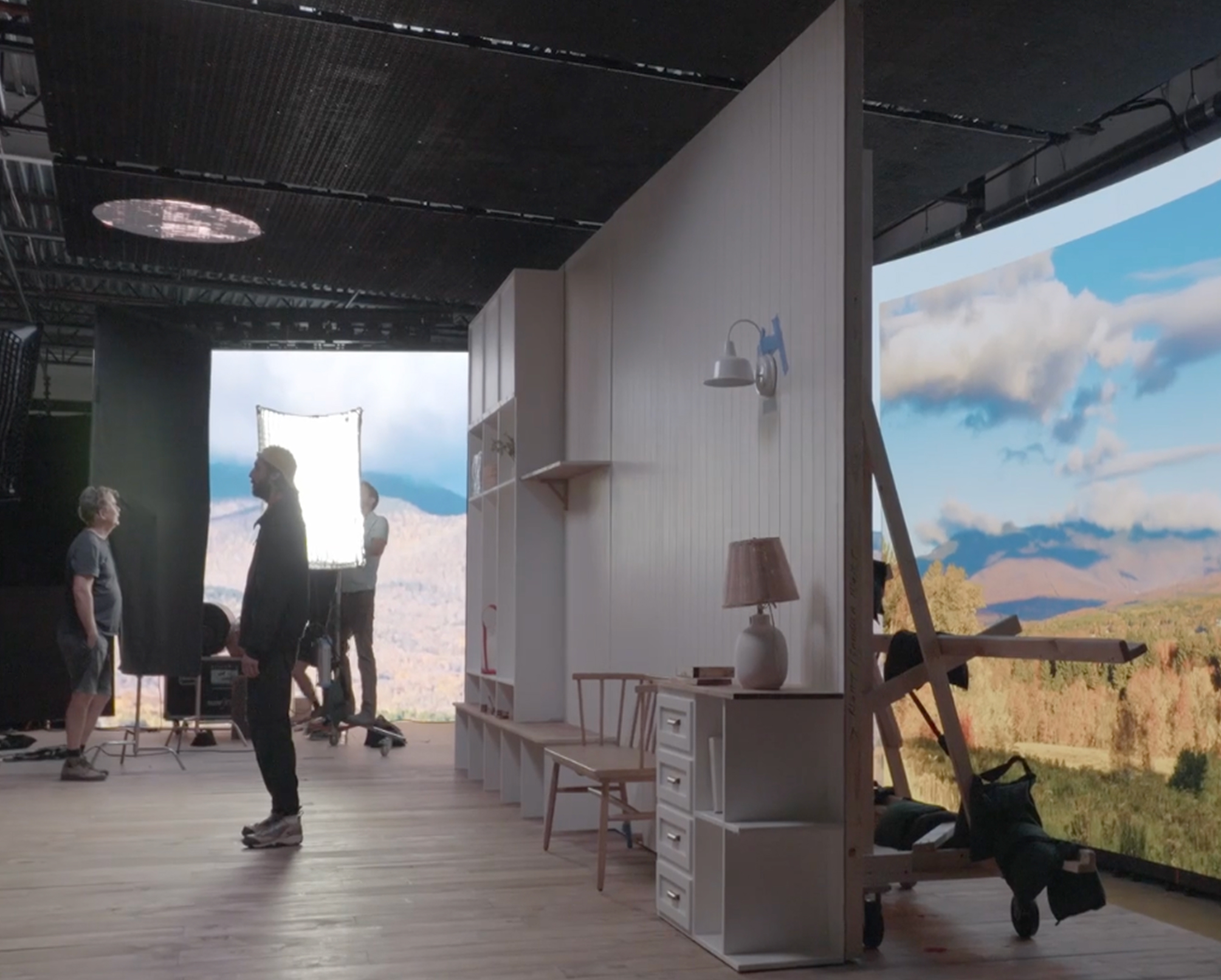 Behind-the-scenes view of a film set featuring a modern interior design with a wall-mounted lamp and a wooden chair. Crew members are adjusting lighting equipment and preparing for a shoot, with a scenic mountain backdrop visible through a large curved screen. The setting showcases a blend of practical elements and advanced technology in film production.