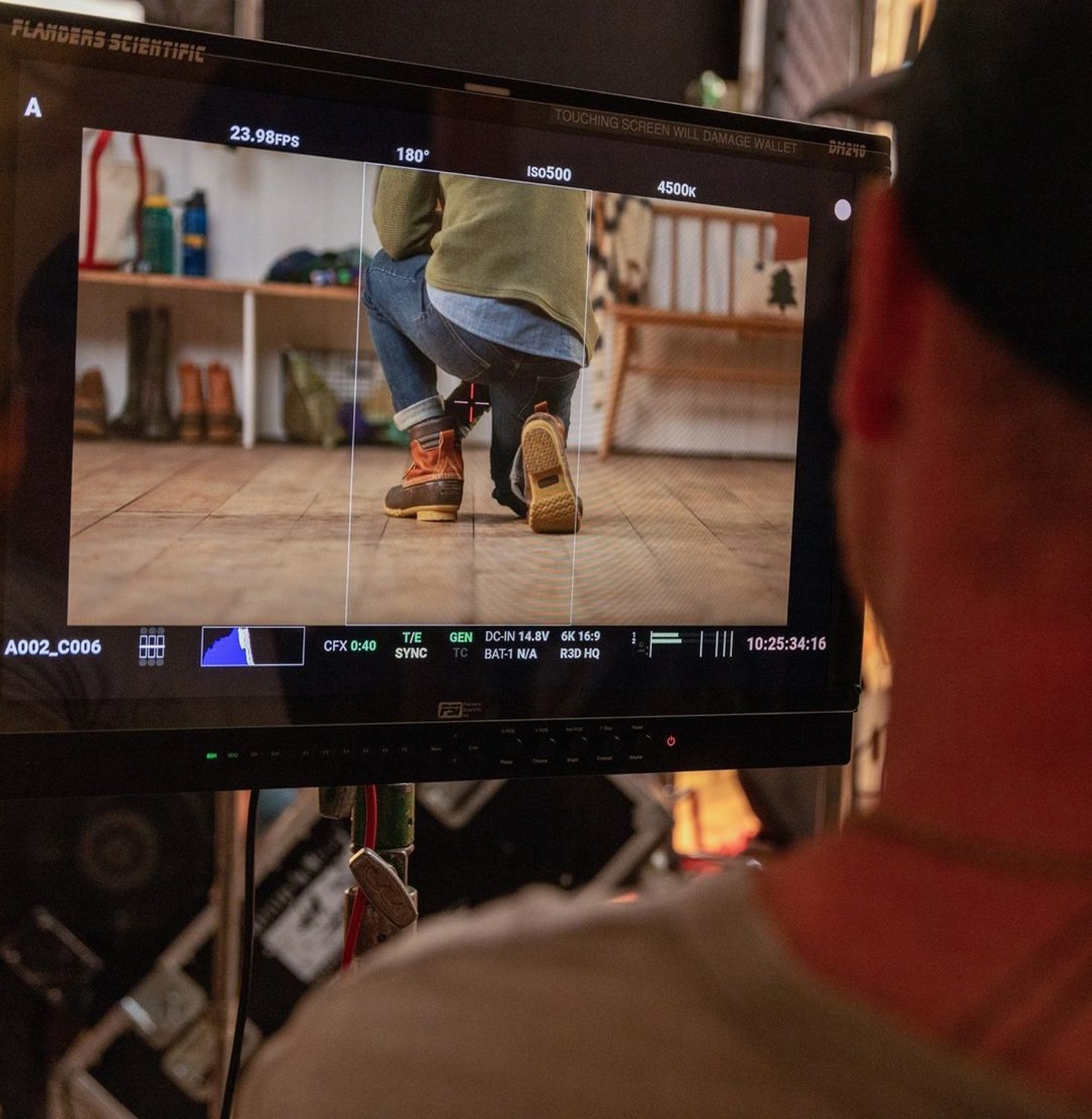 Close-up of a monitor displaying a video shot of a person kneeling in a studio setting, showcasing their footwear and jeans. The background includes various items on a shelf, highlighting a creative workspace environment.