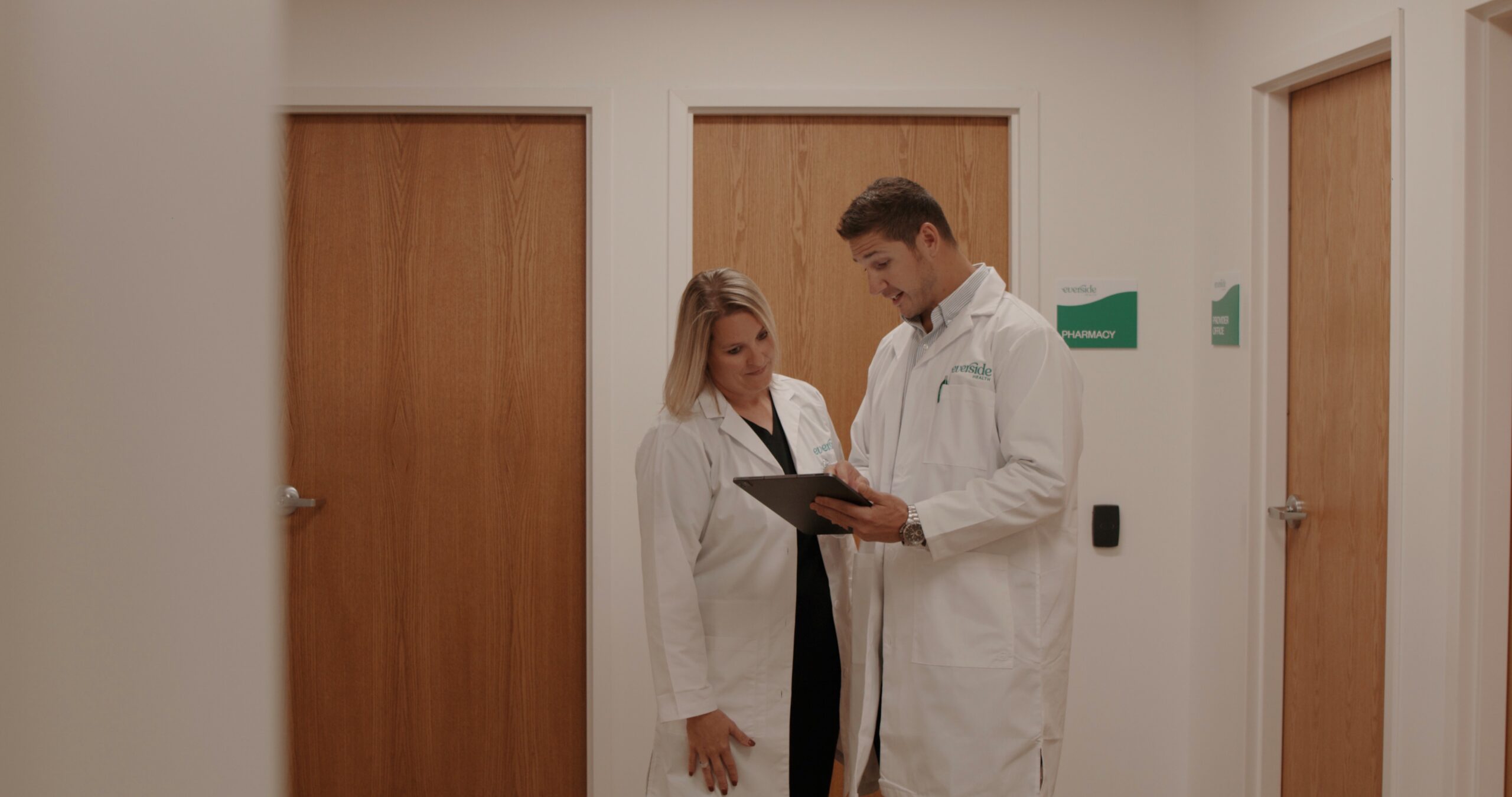 Two medical professionals in white lab coats are discussing information on a tablet in a healthcare setting, with wooden doors labeled "Pharmacy" and "Private Office" in the background.