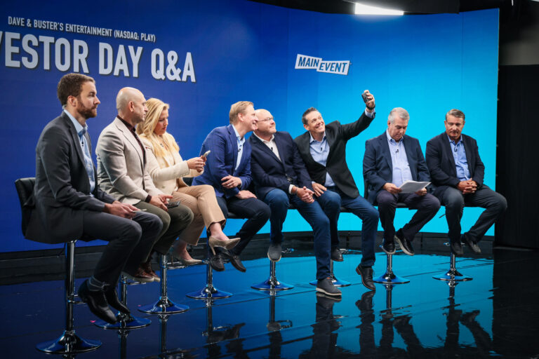 Panel discussion during Dave & Buster's Investor Day Q&A event, featuring executives seated on stools against a blue backdrop with the Main Event logo, engaging with investors and discussing company strategies.