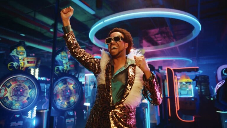 A man in a sparkling outfit celebrates joyfully in a vibrant arcade filled with colorful neon lights and game machines, embodying a fun and energetic atmosphere.