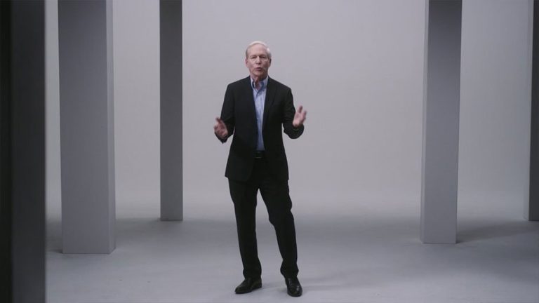 A man in a black suit stands confidently in a minimalist studio setting with gray pillars, gesturing as he speaks, conveying a professional and engaging presence.