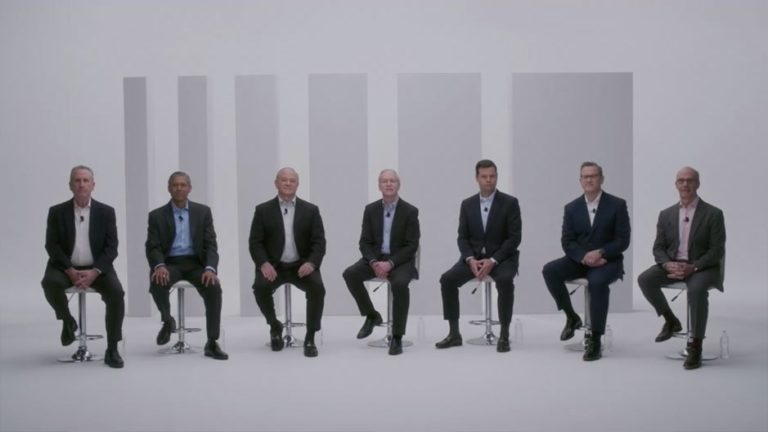 A group of seven business professionals seated on stools in a minimalistic studio setting, wearing suits and microphones, engaged in a panel discussion. The background features simple geometric shapes, creating a modern and clean aesthetic.