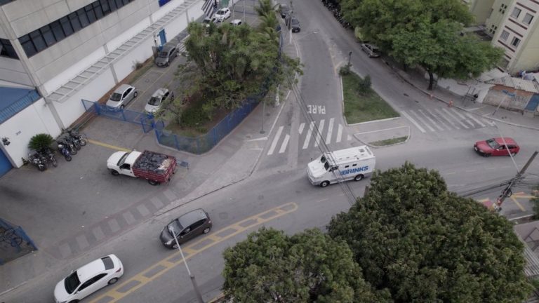 Aerial view of a city intersection featuring a delivery truck, parked cars, and palm trees, with a blue building in the background and a crosswalk visible.