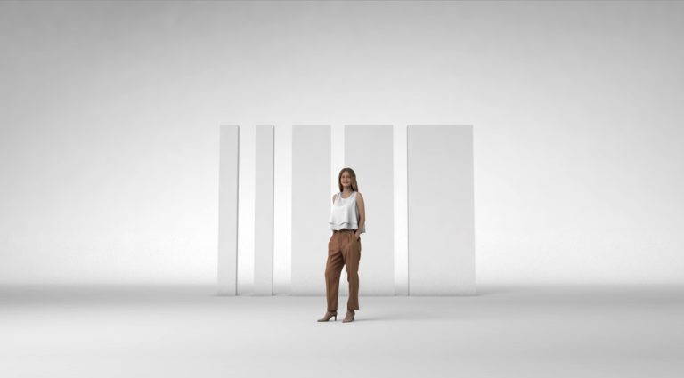 A confident woman stands in a minimalist studio setting, wearing a white blouse and brown pants, with four vertical white panels in the background, highlighting a modern and professional atmosphere.