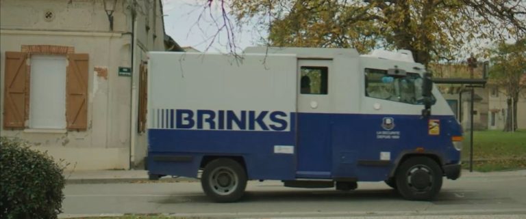 Brinks armored truck driving on a street near a building with closed shutters and foliage in the background.