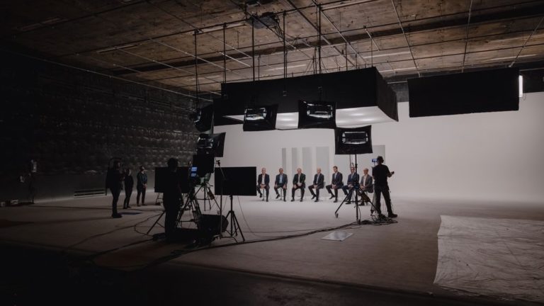 A film studio setup featuring a row of seated individuals in formal attire, with a camera crew and lighting equipment. The backdrop is a plain white wall, and the environment is well-lit, indicating a professional filming session.