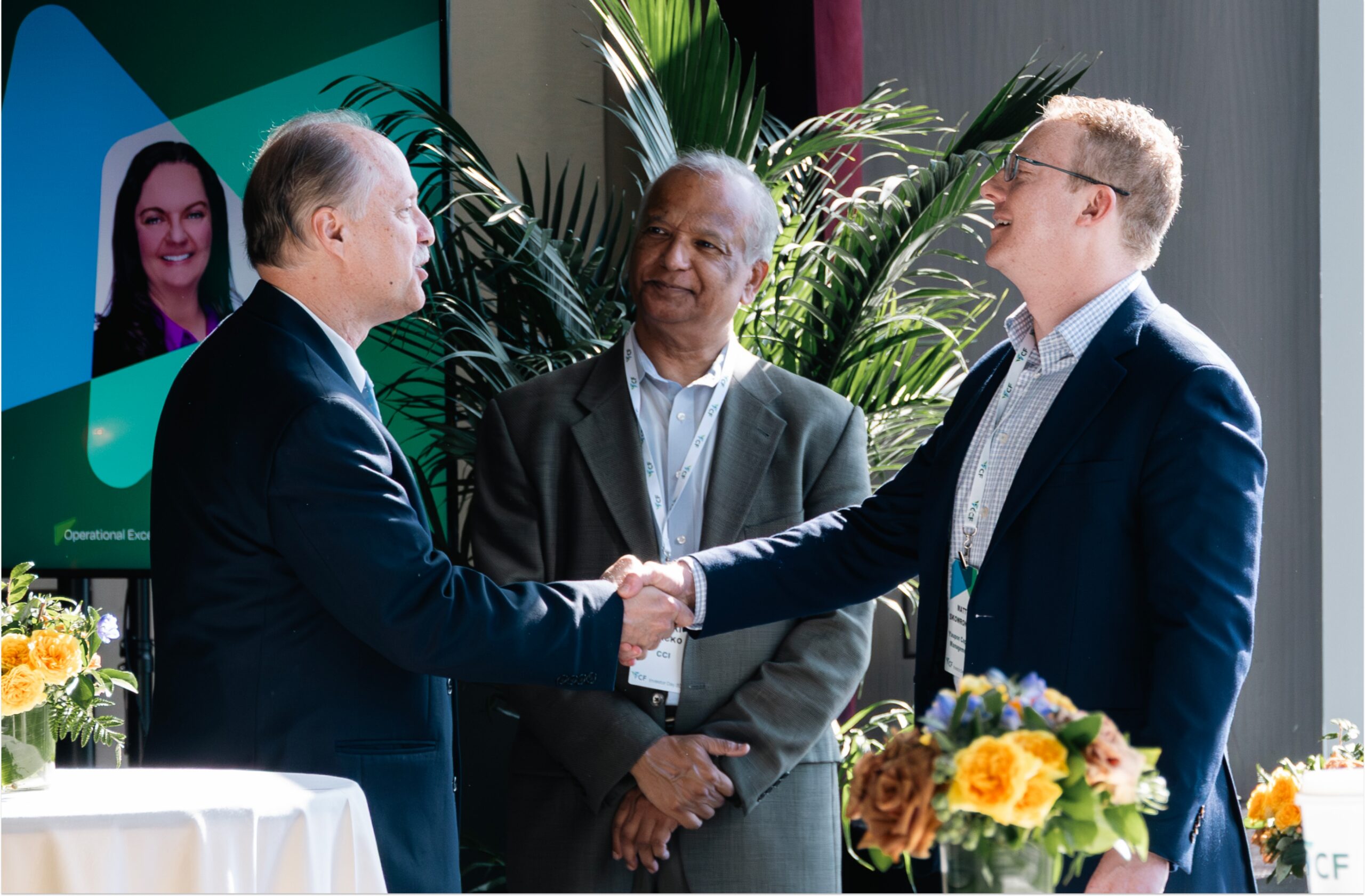Business leaders engaging in a handshake during a corporate event, with a backdrop featuring a presentation screen and greenery, emphasizing collaboration and networking in a professional setting.