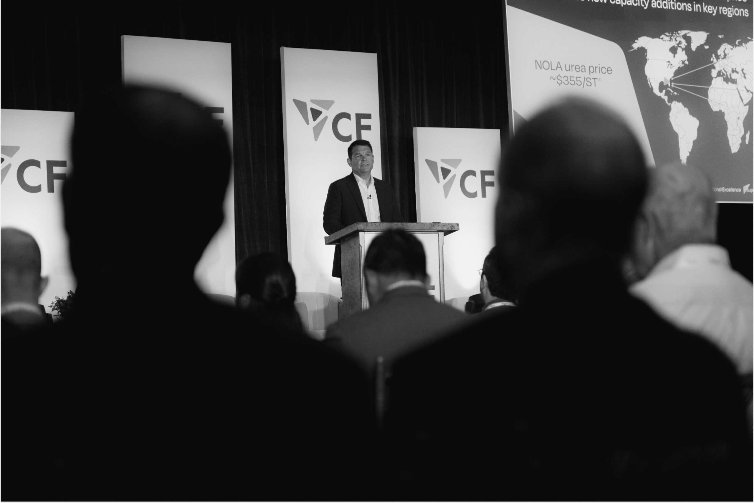 Speaker presenting at a conference with CF branding, discussing market trends and pricing, while audience members attentively listen in the foreground.