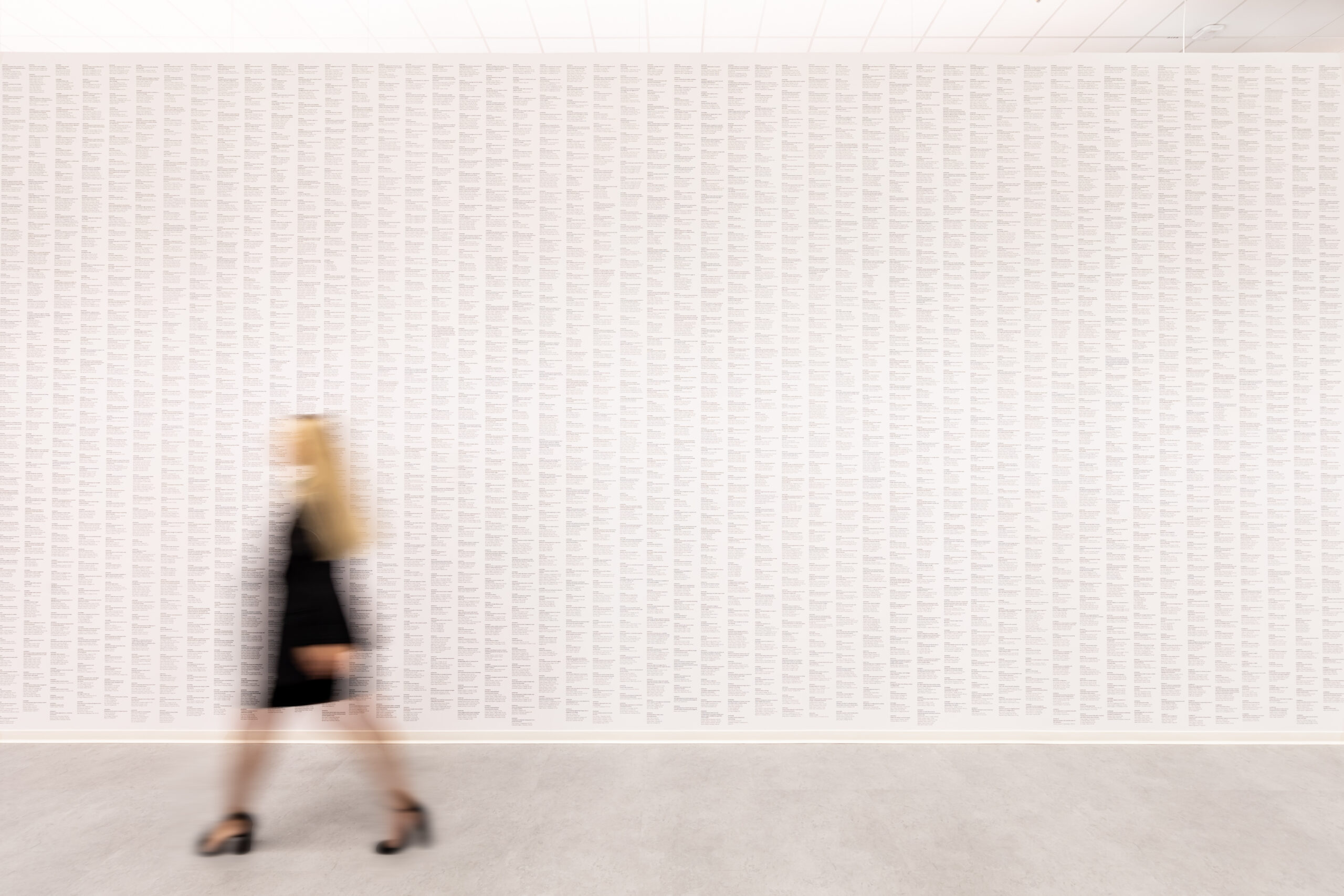 A blurred figure in a black dress walks past a white wall covered with printed text, creating a dynamic contrast in a minimalist art space.