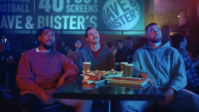 Three friends enjoying food and drinks while watching a sports game at Dave & Buster's, surrounded by a lively crowd and large screens displaying the event.