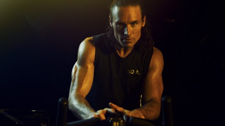 Male fitness instructor in a dark studio, focused on a spinning bike, showcasing muscular arms and intense concentration. The shirt features the word "SOUL," emphasizing a high-energy workout environment.
