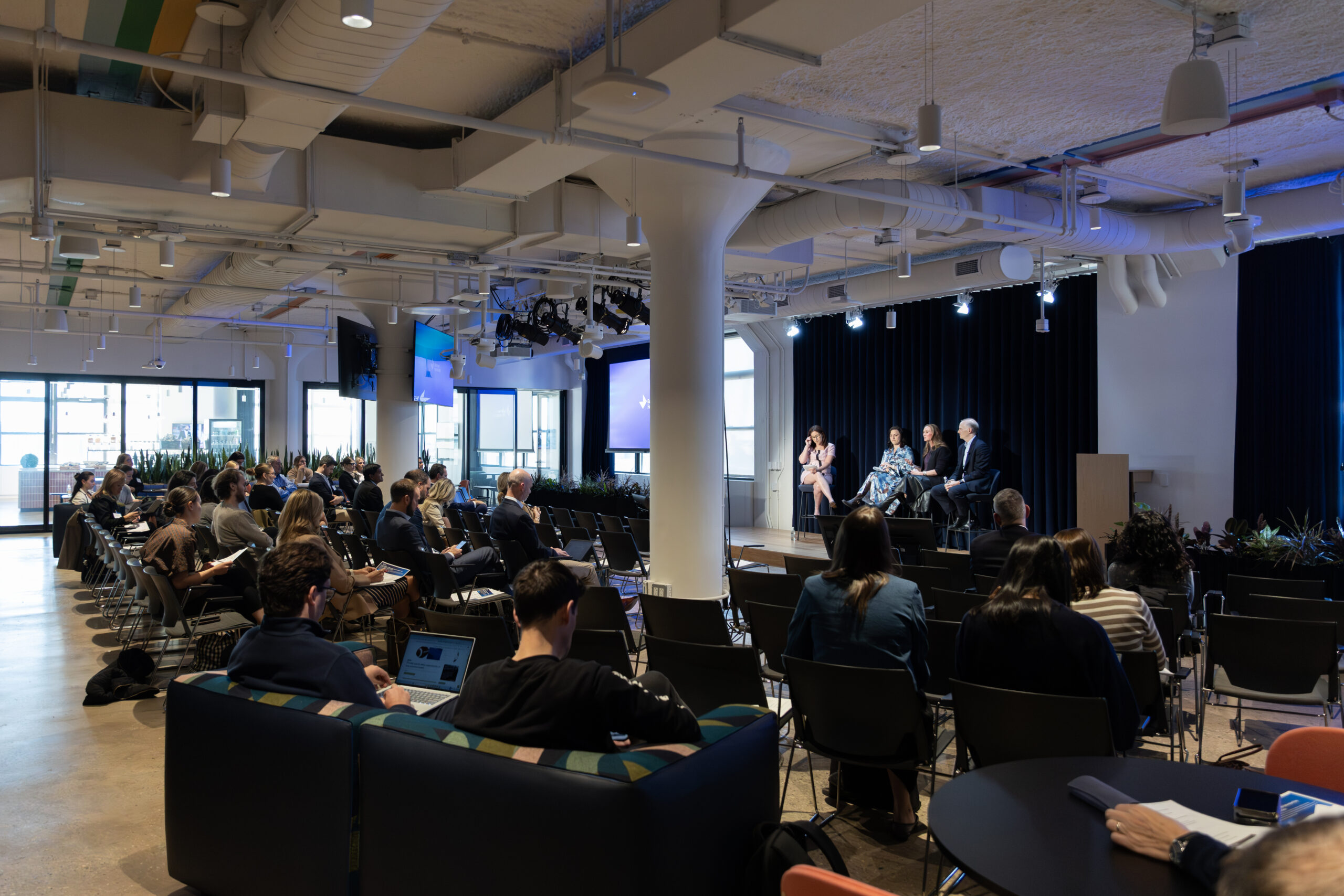 Audience attending a panel discussion in a modern conference space, featuring speakers on stage and attendees seated in rows, with large windows and greenery in the background.