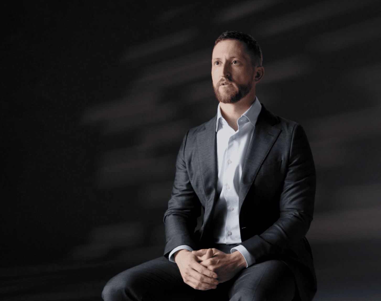 Business professional sitting on a chair, wearing a dark suit and light blue shirt, against a dark background with soft lighting, conveying a serious and contemplative expression.